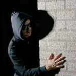 A man in a hoodie hides in the shadows against a brick wall, creating a mysterious mood.