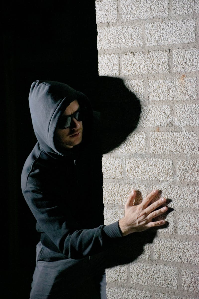 A man in a hoodie hides in the shadows against a brick wall, creating a mysterious mood.