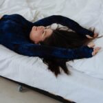 A woman with long hair stretches and relaxes on a bed wearing a blue top, enjoying a peaceful moment indoors.
