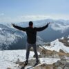A man stands on a snowy mountain peak with arms raised, embracing the breathtaking view.