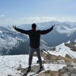 A man stands on a snowy mountain peak with arms raised, embracing the breathtaking view.