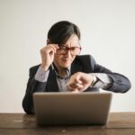 Young frowning man in suit and glasses looking at wristwatch while waiting for appointment sitting at desk with laptop