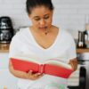 A woman reads a red book in a contemporary kitchen setting, showcasing a cozy lifestyle.