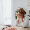 A woman sitting at a desk, overwhelmed by financial stress, with bills and calculator in a bright room.