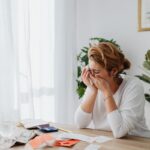 A woman sitting at a desk, overwhelmed by financial stress, with bills and calculator in a bright room.