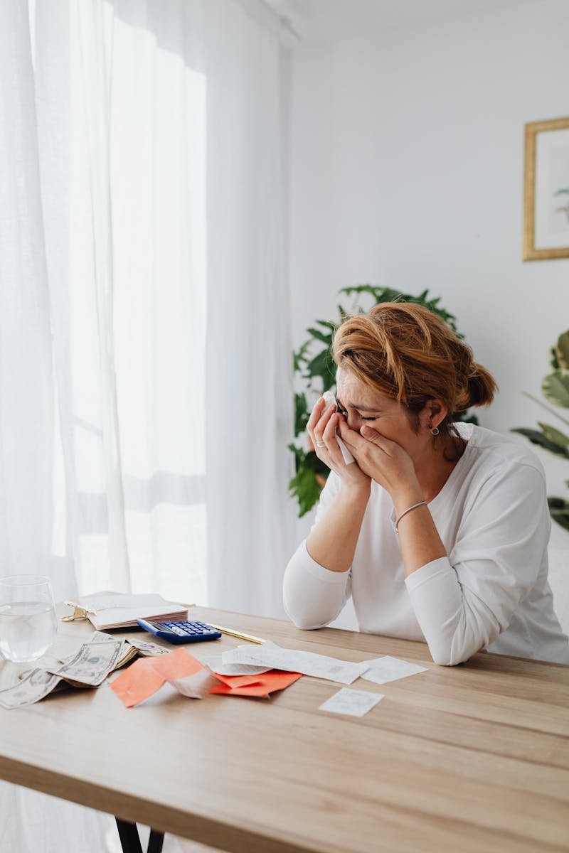 A woman sitting at a desk, overwhelmed by financial stress, with bills and calculator in a bright room.