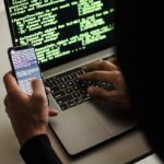 A hooded figure engaged in hacking using a laptop and smartphone in low light.